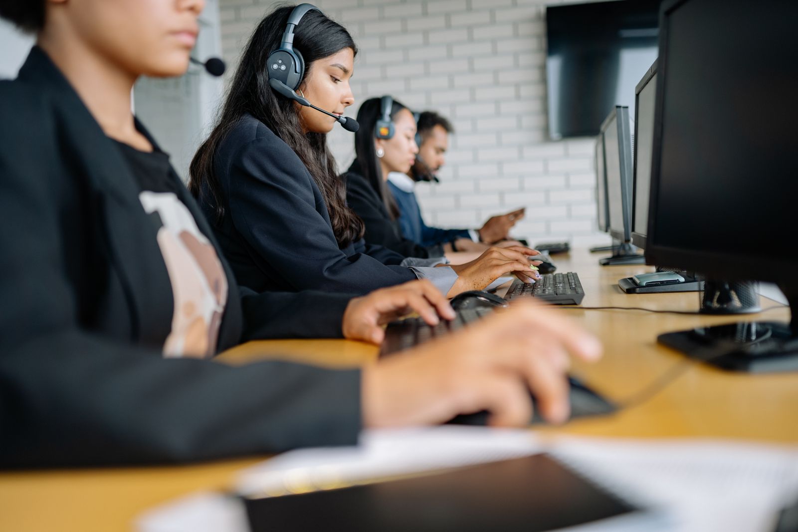 A row of contact centre agents on headsets typing at monitors along a shared desk, seen from the side